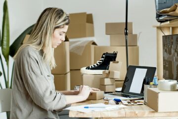 Small e-commerce business owner packing orders at a desk with laptop and boxes, managing online sales and shipping.