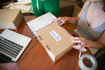 Two people packing online orders at a small business desk with a laptop and shipping materials.