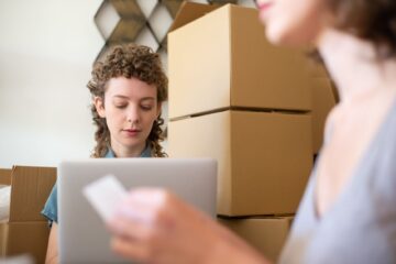 Small ecommerce business owner packing orders with laptop and cardboard boxes in a compact workspace.