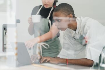 Two cafe staff using a laptop to manage bookings and online orders in a small restaurant.