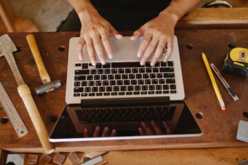 Workshop owner using a laptop at a wooden workbench in a carpentry workshop.